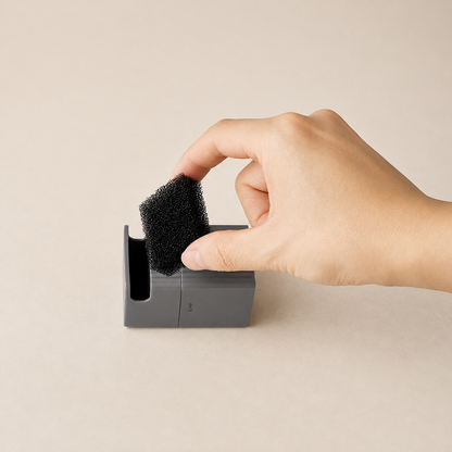 Hand holding a black replacement filter over a grey holder on a beige background