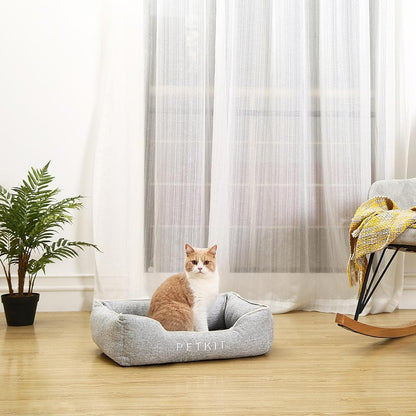 Cat sitting in a gray PETKIT pet bed in a room with curtains and a plant.