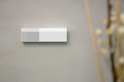 White rectangular smart odour eliminator mounted on a grey wall with a blurred decorative plant in the foreground.