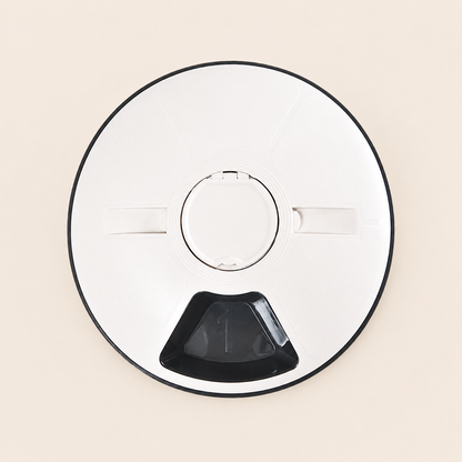 A top-down view of a circular automatic pet feeder with a black rim, a central circular opening, two small side tabs, and a black triangular opening at the bottom, set against a solid light beige background.