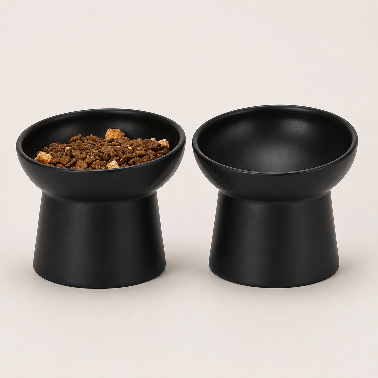 Two matte black pedestal pet bowls on a light beige background, with one bowl filled with brown kibble.